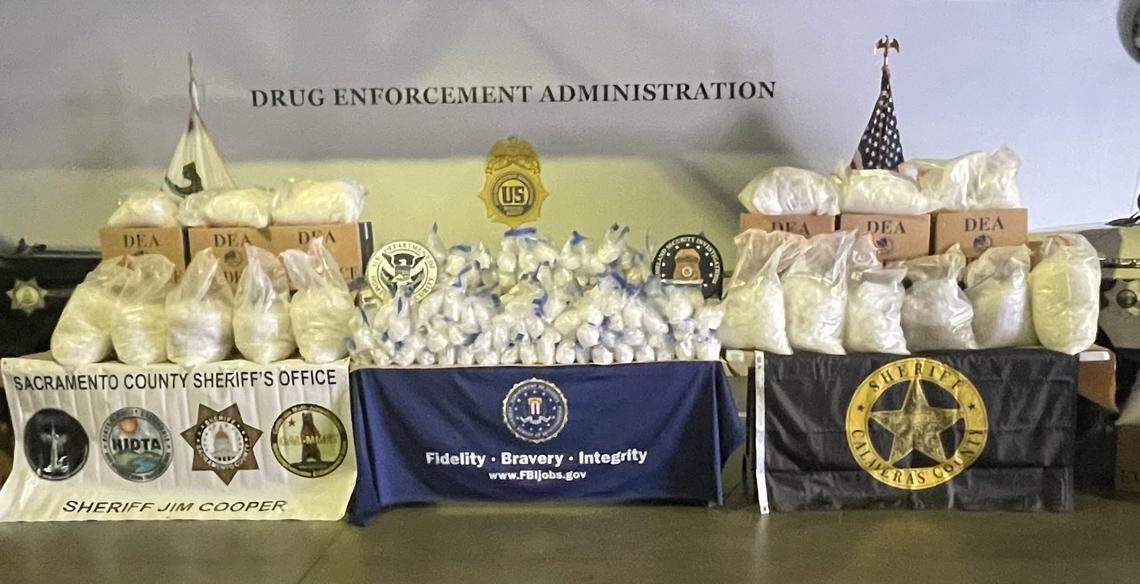 Bags of suspected methamphetamine and boxes labeled as evidence are displayed alongside law enforcement insignia during a multi-agency operation that authorities say resulted in the seizure of roughly 2,700 pounds of methamphetamine.