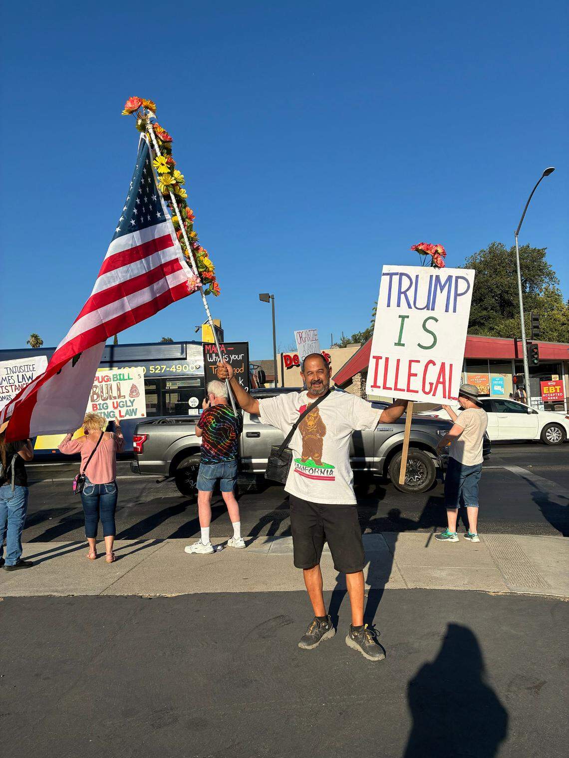 A man in a California flag inspired t-shirt holds a sign that reads "Trump is illegal" in one hand and a double-sided flag in the other. One side is the US flag, the other the California state flag. 