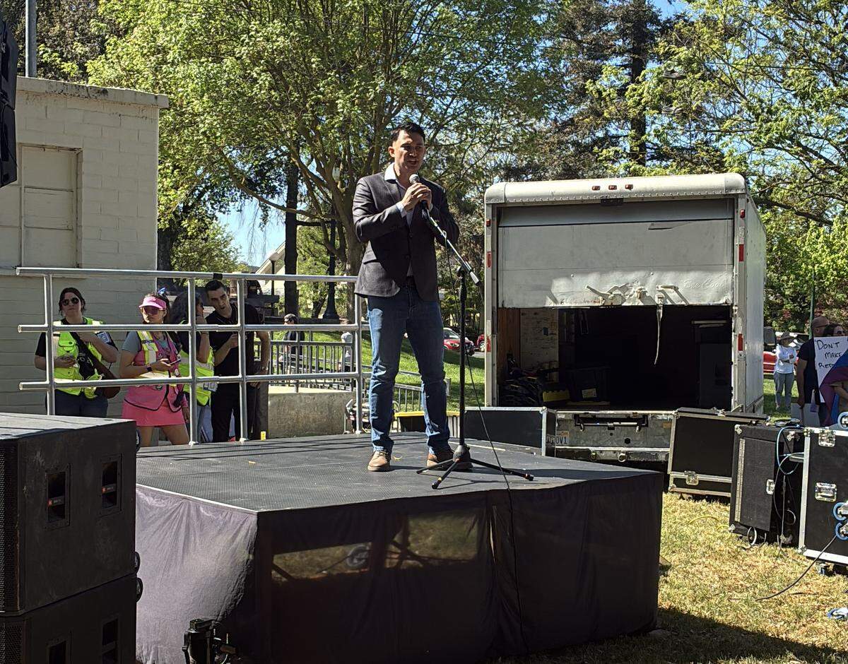 A man in his thirties in a suit stands at a microphone on a stage in Graceada Park.