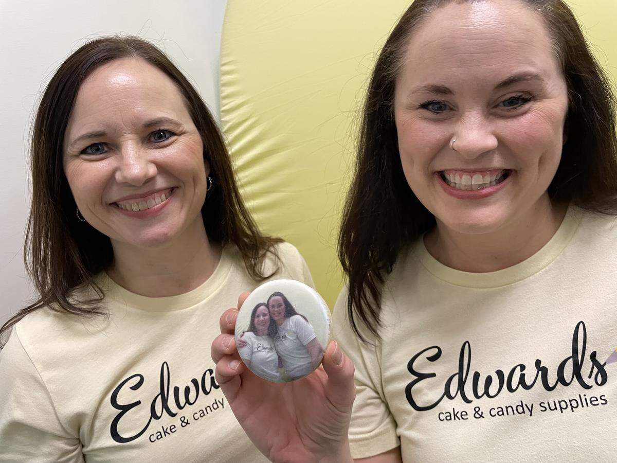 Sisters Emily Damante and April Brown, the co-owners and operators of Edwards Cake and Candy Supplies with a sugar cookie from their Cookie Photobooth. The machine prints photos onto sugar cookies in an instant.