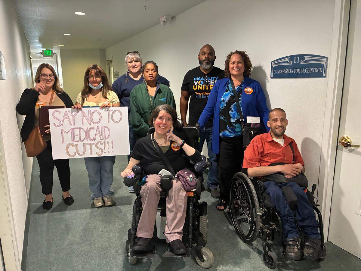 Two people in whellchairs and six others behind them stand outside of McClintock's closed office door. One member holds a sign that reads "Say no to MediCaid cuts!!!"