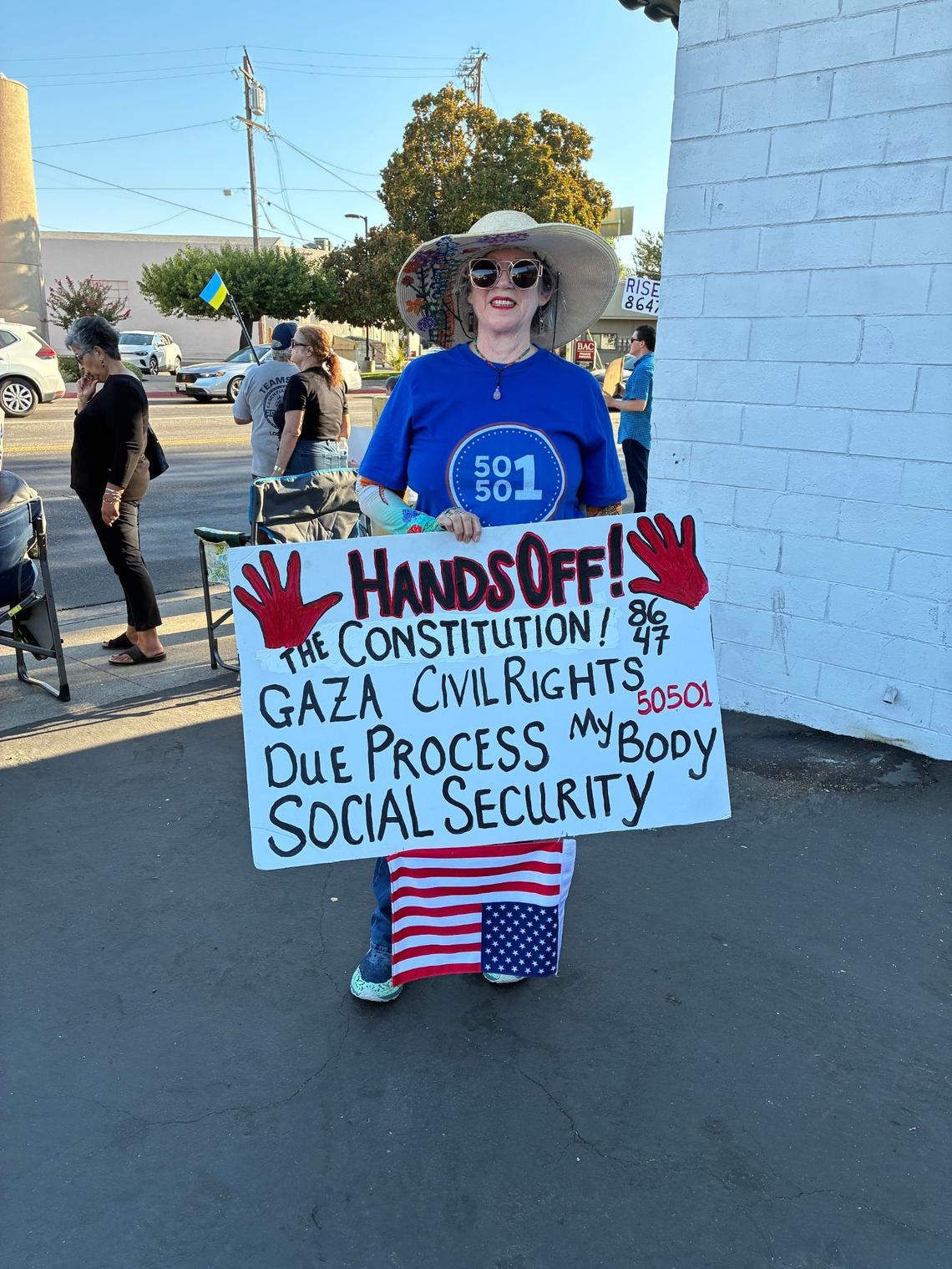 A woman wearing sunglasses and a sun hat that has been embellished with flowers under the brim hold a sign by her feet that reads "Hands Off the constitution, Gaza, my body, due process, socials security, 50501" and "8647"
