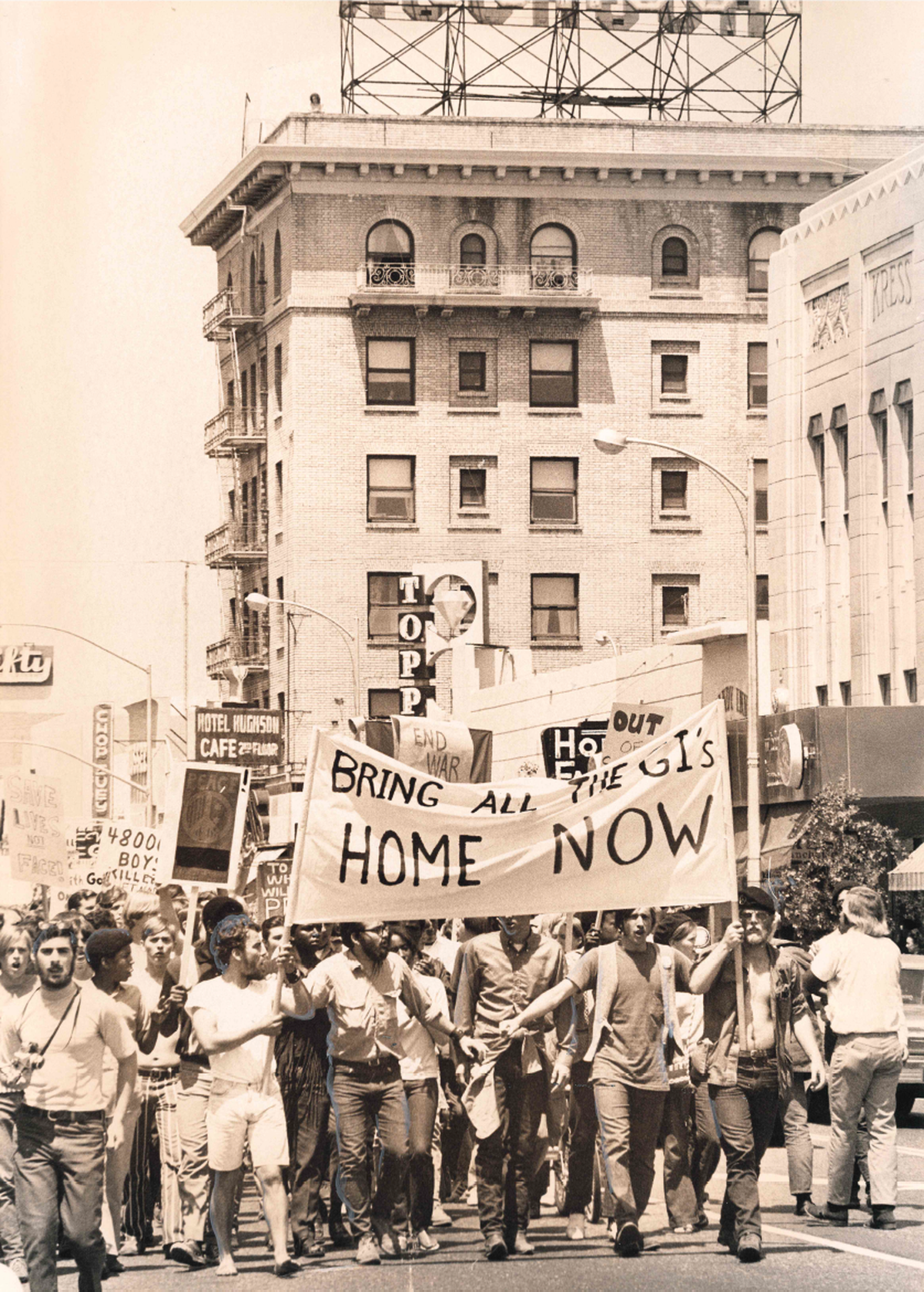A large group of people march down 10th street in downtown Modesto. Several hold up a sign that reads "Bring all GIs home now" to protest the Vietnam War