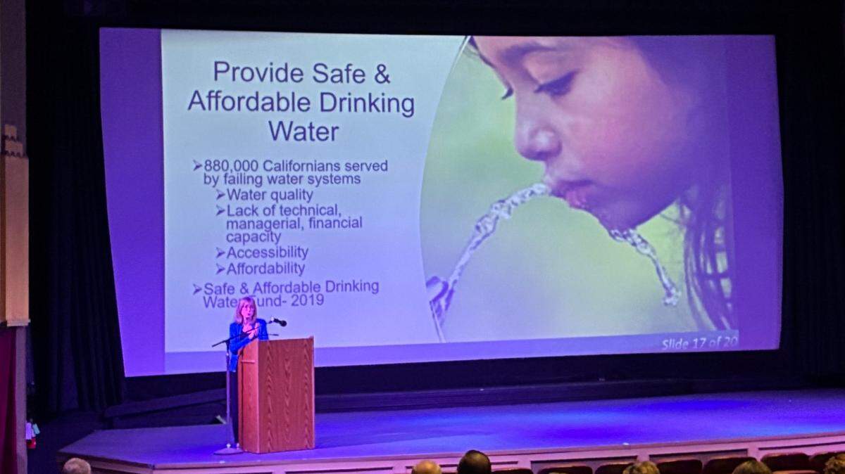 A woman stands at a podium overlooking the State Theater auditorium. Behind her is a slide on the state's plan to provide safe and affordable drinking water.