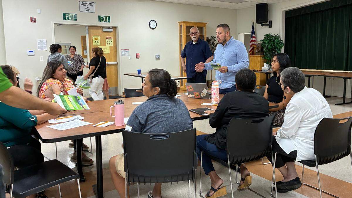 Several people, some sitting, some standing, gather around a table and listen to differing perspectives on a prospective new soccer stadium.
