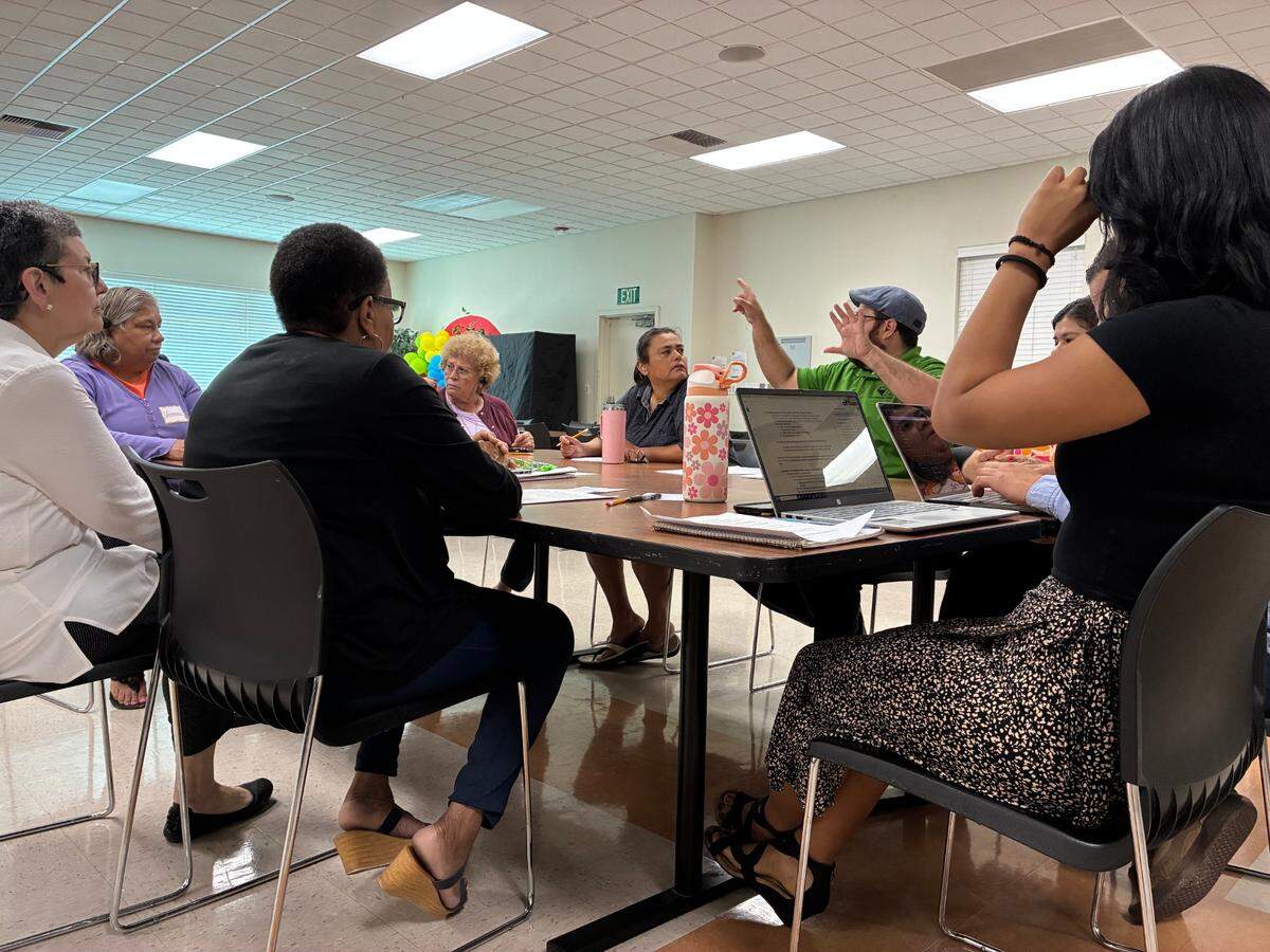 Community members and advocates talk through a community issue around a brown table. Edgar Garibay, one of the presenters, holds his hands up to emphasize his point.