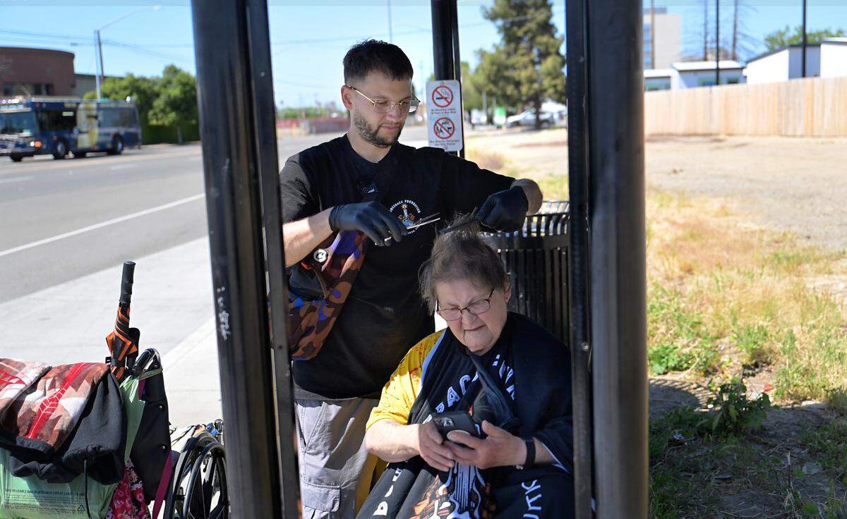Paul “Sammy” Samsin cuts the hair of Katherine Root at a bus stop on Ninth Street in Modesto, Tuesday, April 8, 2025. Sammy is a licensed barber and formerly homeless, he has been giving back by cutting hair of homeless people.