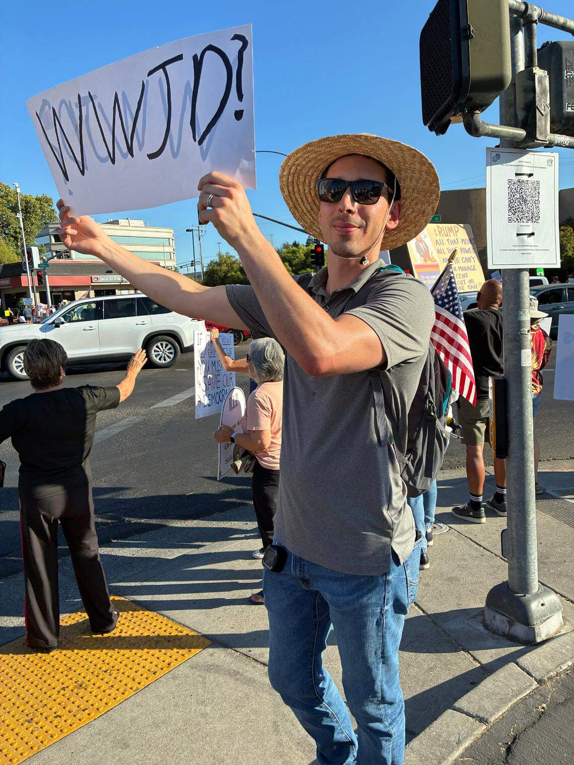 A man wearing a sunhat and sunglasses holds a sign that reads "WWJD?"