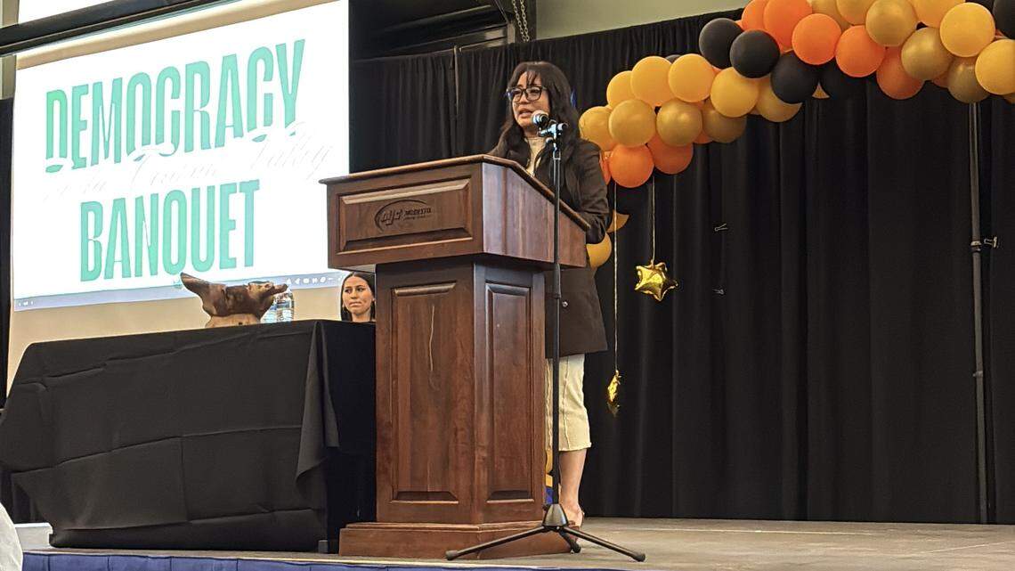 A woman stands at the podium, behind her is a balloon arch and a screen that reads “Democracy In The Central Valley Banquet.”