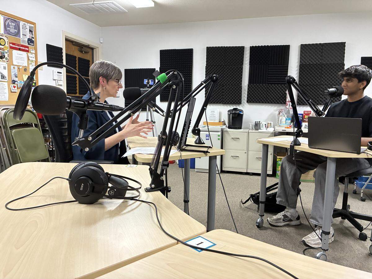 A teenager sits in a chair with a desk and microphone in front of him across from a woman who sits in her own chair with a mic in front of her.