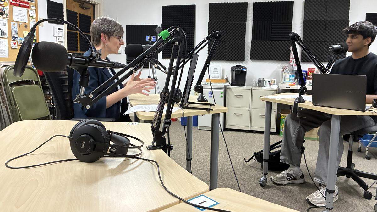 A teenager sits in a chair with a desk and microphone in front of him across from a woman who sits in her own chair with a mic in front of her.