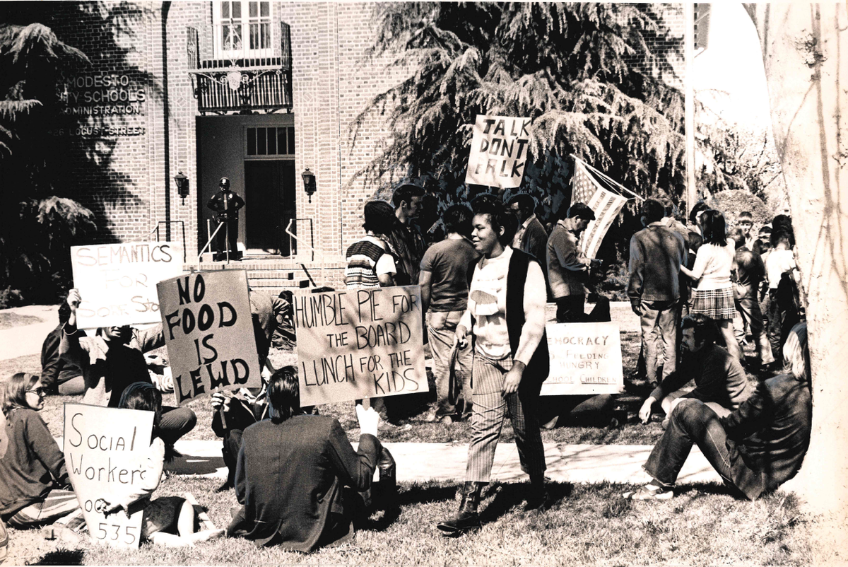 Protesters sit outside of Modesto City Schools and hold signs that read "Humble pie for the board, lunch for the kids" and "no food is lewd"