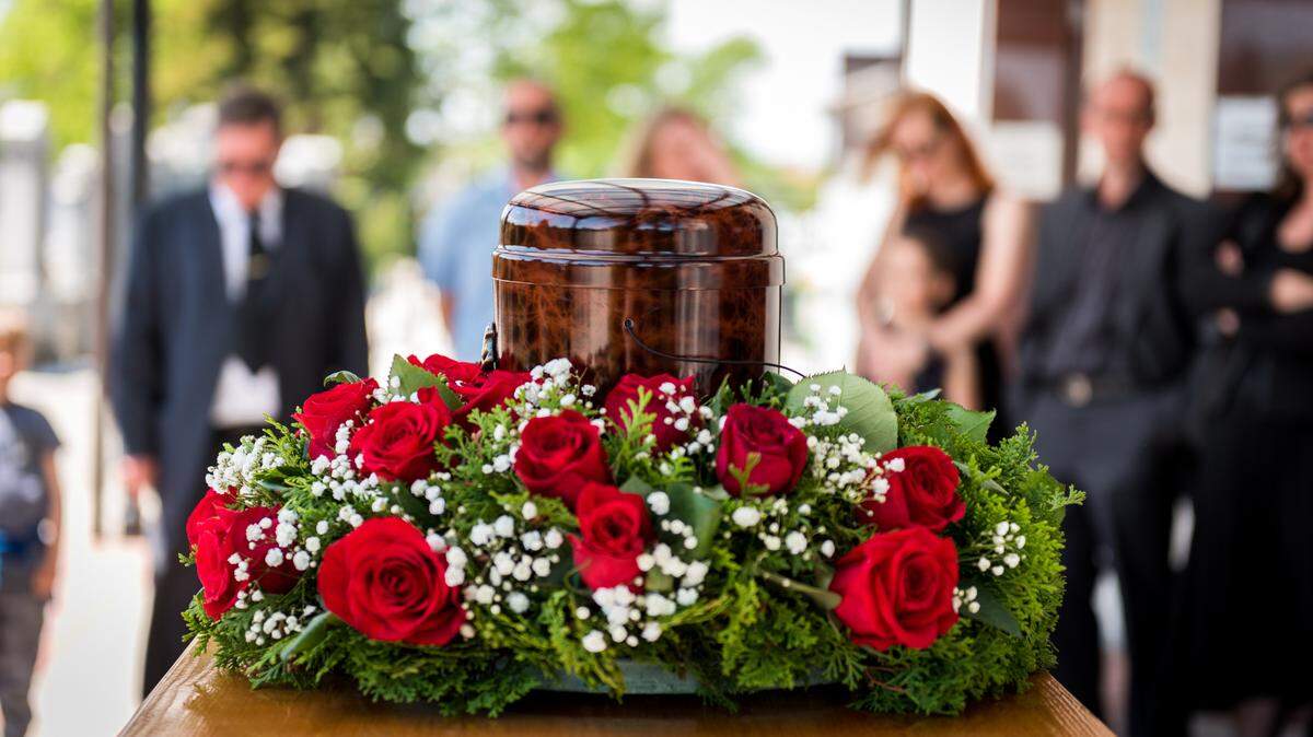 Funerary urn with ashes of dead and flowers at funeral. Burial urn decorated with flowers and people mourning in background at memorial service, sad and grieving last farewell to deceased person.