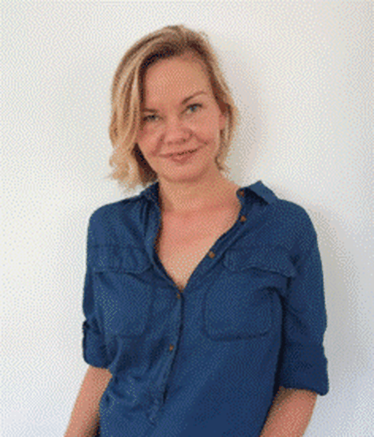 A woman with short blonde hair in a blue button down shirt smiles looking into the camera.