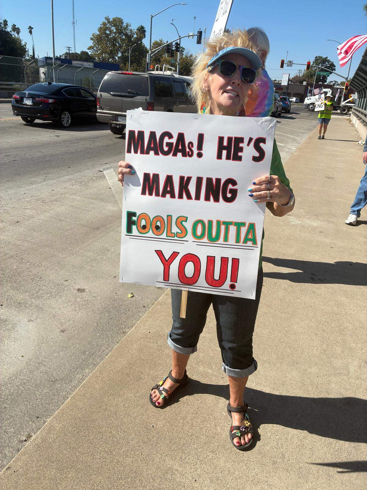 A woman in a visor and sunglasses holds up a sign that reads “MAGAs, he’s making fools outta of you!”