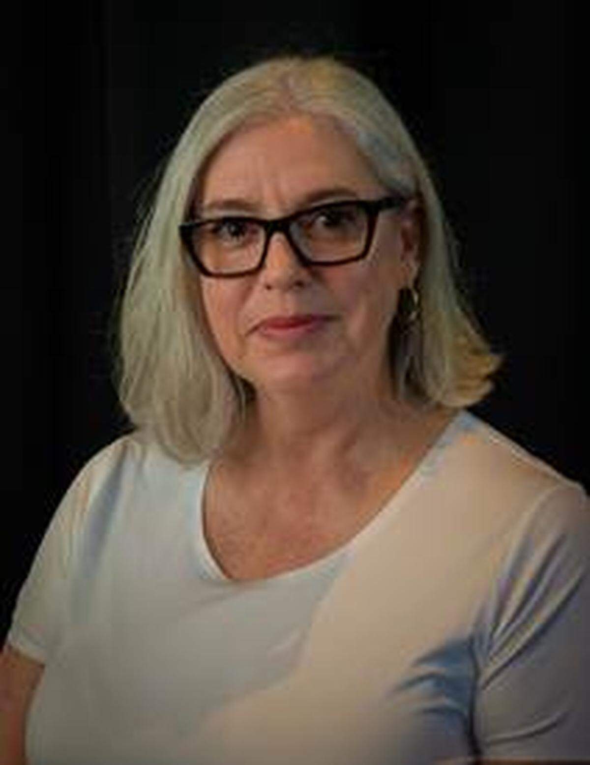 A woman wearing a light gray shirt and black glasses with shoulder length gray hair looks forward towards the camera with a slight smile on her face.