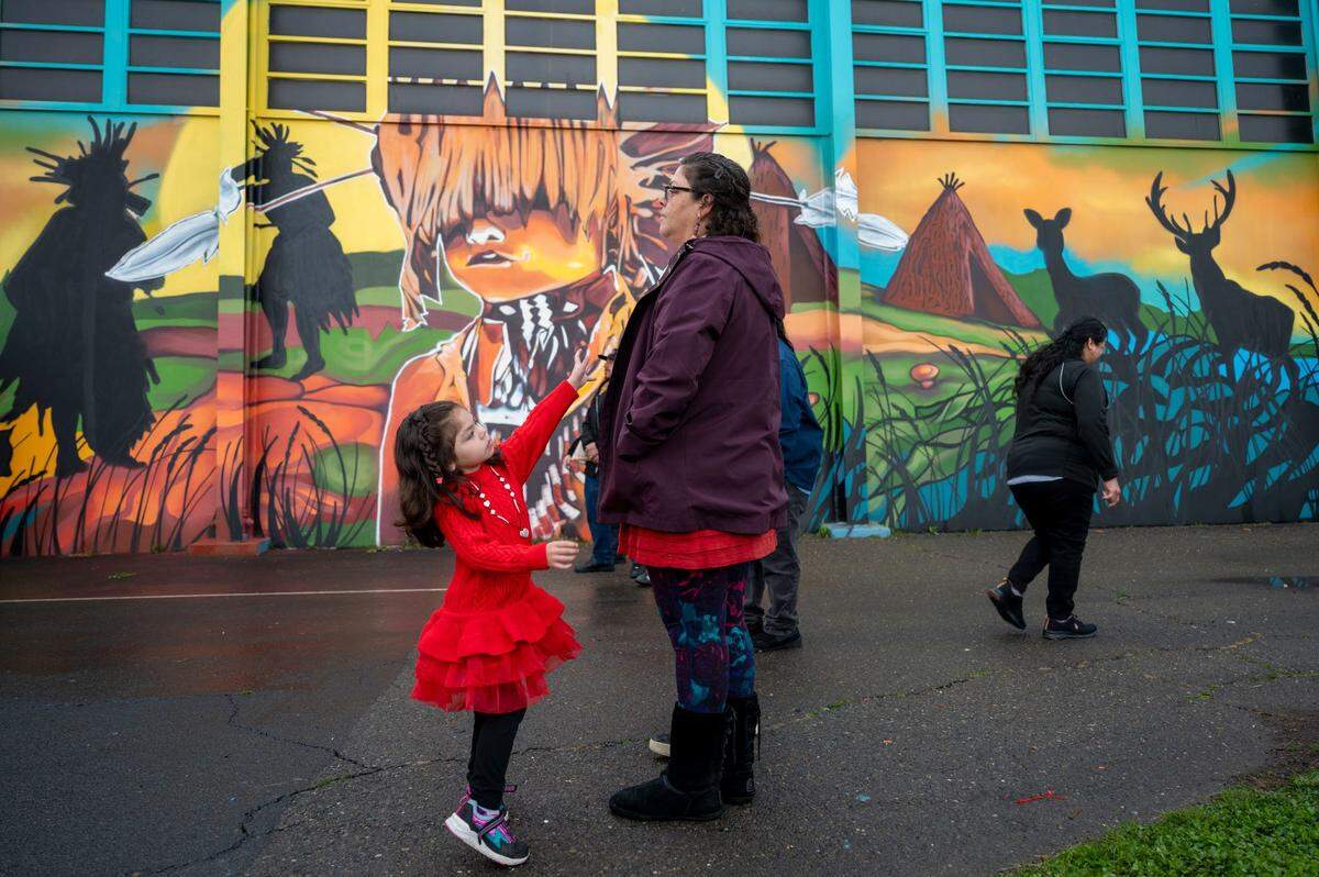 Iris Williams, 4, reaches for her mother Viola Williams, a tribal citizen of the Wilton Rancheria, during the unveiling of a mural on the outside of the gym at Miwok Middle School on Saturday, March 30, 2024, that celebrates its name change from Sutter Middle School.