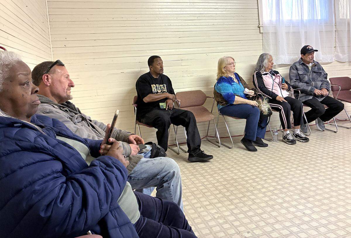 Six people sit in pink chairs along the edge of the room looking towards the center.