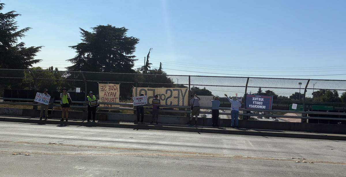 Eight people, two wearing neon yellow vests, stan on the L street overpass holding signs and looking towards the main road or the freeway.