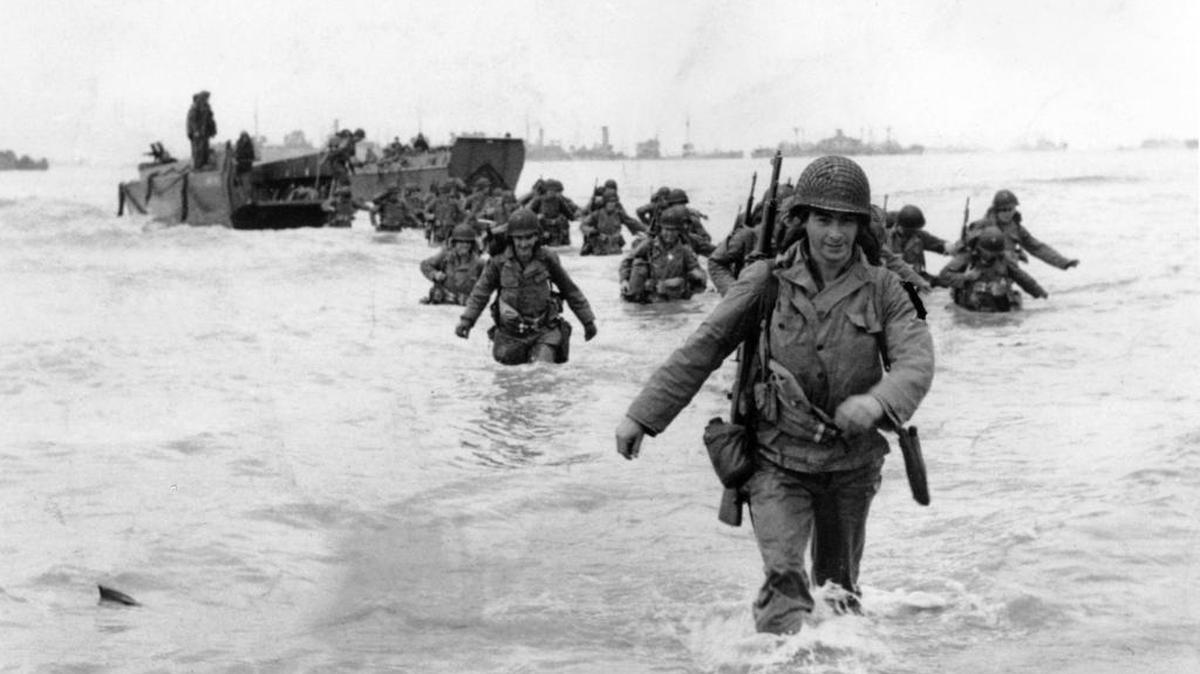 U.S. infantrymen wade through the surf as they land at Normandy in the days following the allies’ June 1944 invasion of occupied France. An allied ship loaded with supplies and reinforcements waits on the horizon.