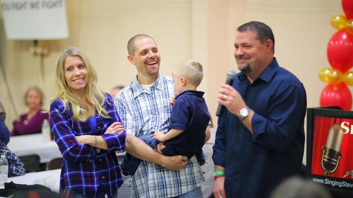 Kirk Clifton (right) addresses the crowd at a dinner and auction benefitting his brother Kraig (center), his wife Alissa (left) and their four children.